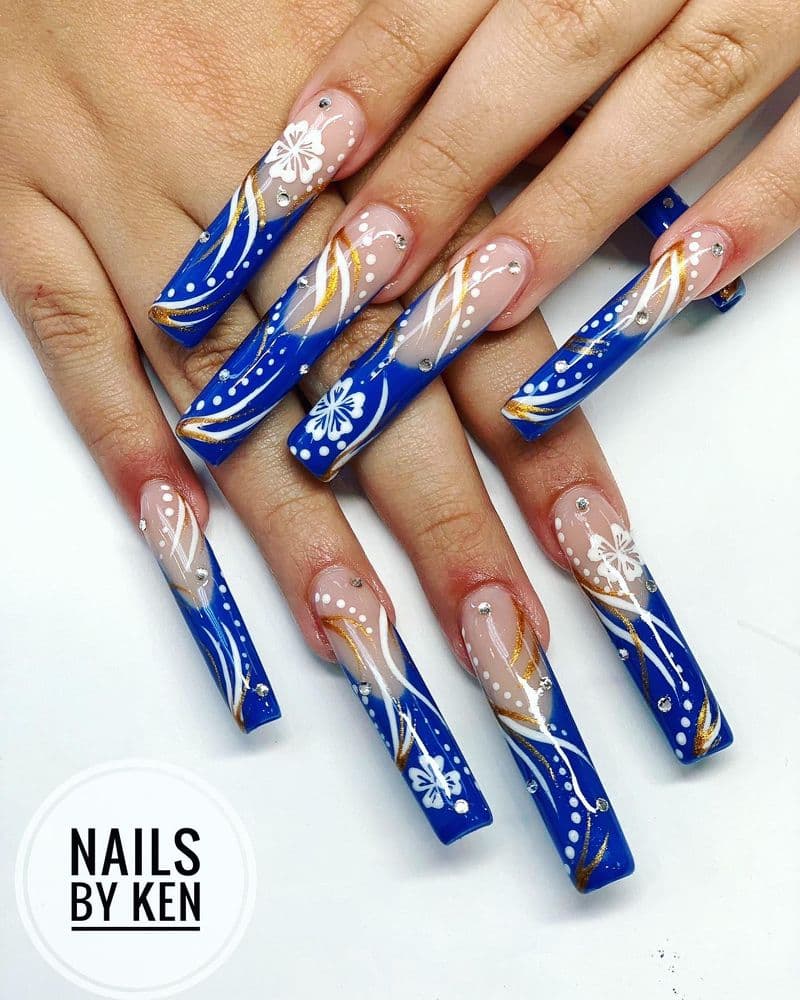 Long square nails with royal blue tips, white floral designs, gold swirls and crystal rhinestones by Nails by Ken
